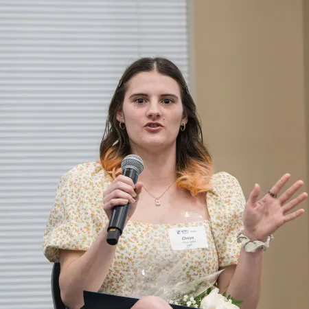 Young woman in floral dress speaking into microphone while seated, holding flowers and folder at event.
