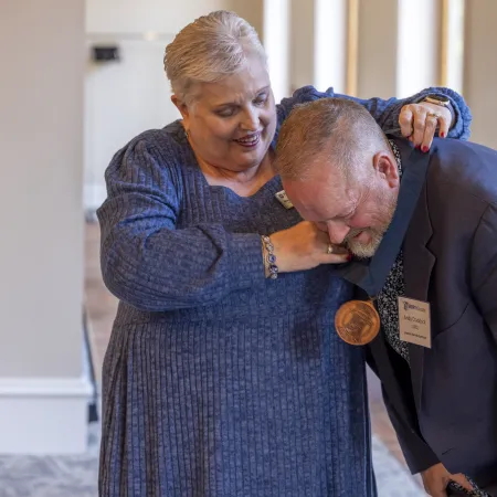 Older woman places a bronze medal around a smiling older man's neck in a warmly lit room.