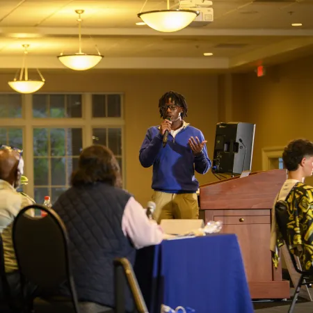 Young man in blue sweater giving a presentation to seated audience in a well-lit conference room.