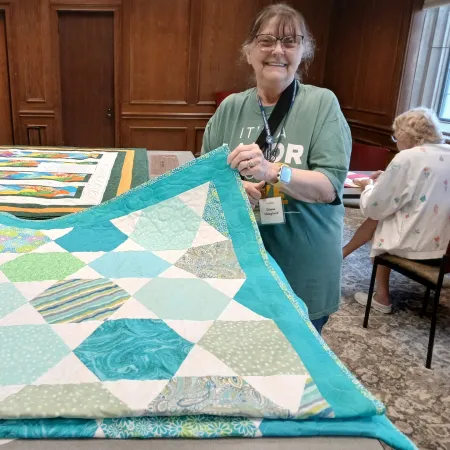 Smiling woman holding a colorful blue and green patchwork quilt inside a wood-paneled room with another quilt in the background