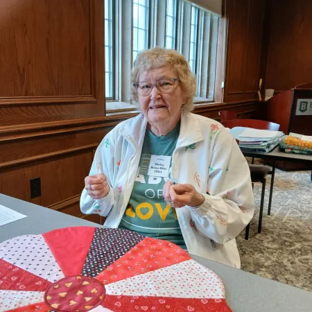 Elderly woman sewing a colorful quilt at a table in a wood-paneled room with large windows.