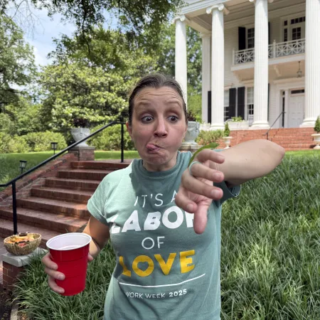 Woman making a funny face holding a red cup in front of a large white-columned house with brick steps.