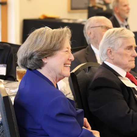 Elderly man in suit with red tie and smiling woman in blue blazer seated at formal event.