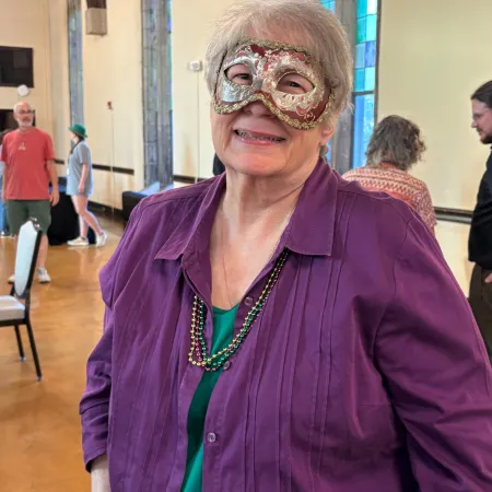 Older woman wearing a decorative mask and purple shirt indoors with stained glass windows in the background