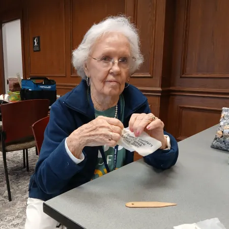 Elderly woman sitting at a table crafting with fabric in a cozy wood-paneled room.