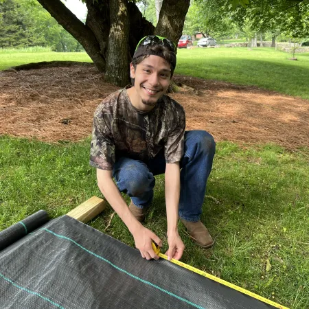 Young man in camouflage shirt measures landscape fabric outdoors on grass near large tree on sunny day