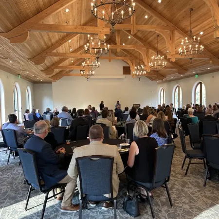 Business conference in a spacious hall with wooden ceiling, chandeliers, and attendees seated at round tables.