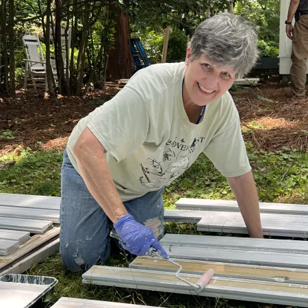 Smiling woman wearing gloves painting wooden planks white outdoors on grass during a sunny day.