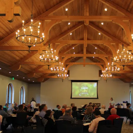 Spacious banquet hall with wooden vaulted ceiling, chandeliers, and attendees seated for a presentation.