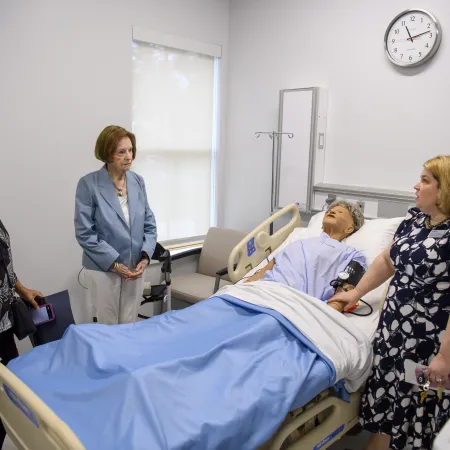 Three women stand around a hospital bed with a medical mannequin used for training in a clinical room.