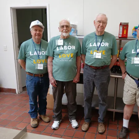 Four senior men standing indoors wearing matching green 'Labor of Love' t-shirts during work week event.