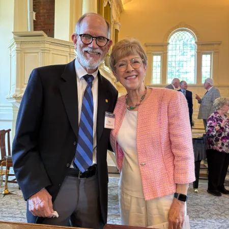 Smiling elderly man in suit with blue striped tie and woman in pink jacket posing in elegant room with arched windows.