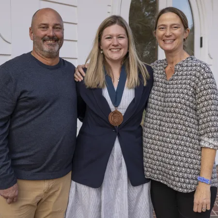 Three smiling adults standing outdoors, center woman wearing a medal around her neck, posing for a photo