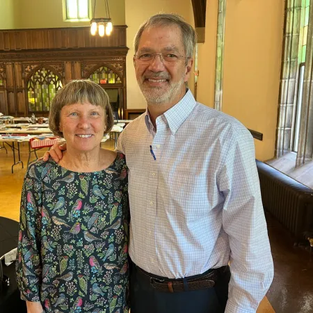 Smiling older couple standing in a large room with tables and wooden paneling in the background.
