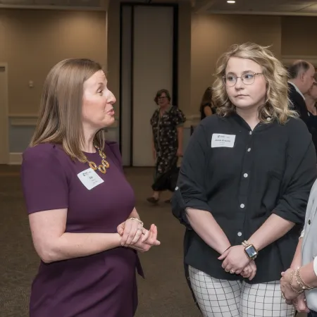 Three women in conversation at an indoor event, wearing business casual attire with name tags.
