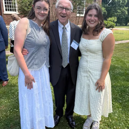 Three people standing outside on grass smiling, two women in dresses and a man in a suit with tie and glasses.