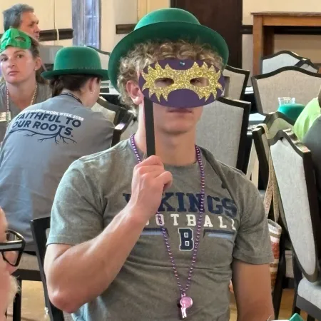 Young man wearing a green hat and holding a glittery Mardi Gras mask on a stick at a festive indoor event.
