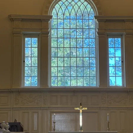 Large arched stained glass window with intricate framing behind a white ornate altar and floral arrangement inside a church.