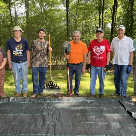 Group of seven volunteers standing outdoors with gardening tools on a sunny day preparing yard work area.