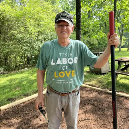 Smiling man in a camouflage cap and green shirt stands in a garden holding a rake and shovel with trees and picnic table behind.