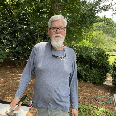 Older man with white beard and glasses, holding a hammer outdoors in a garden setting with greenery.