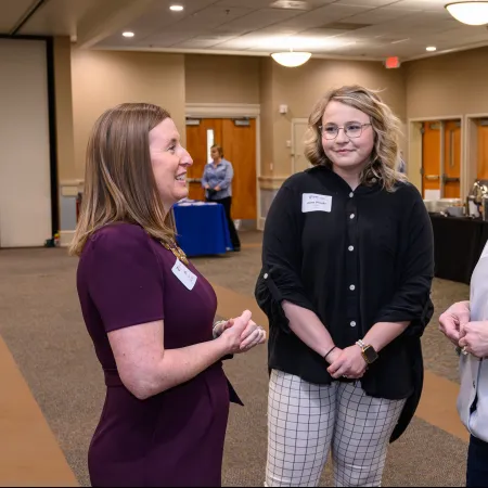 Three women engaged in conversation at a professional indoor networking event with name tags visible.