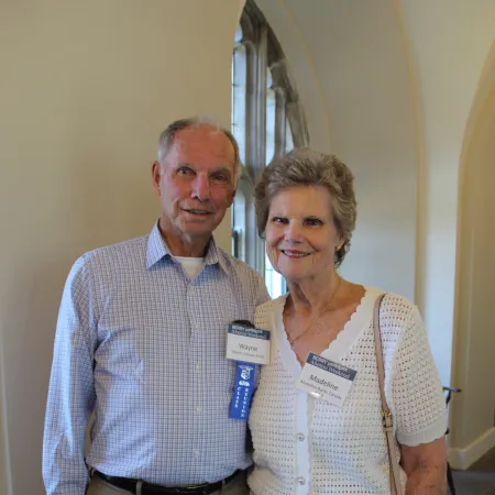 Elderly couple with name tags posing indoors in casual attire with a window in the background
