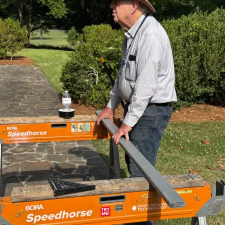 Man in a hat demonstrating the orange Speedhorse metal bending machine outdoors on a stone pathway.