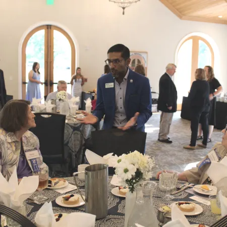 People socializing and dining at a formal indoor event with round tables and arched windows.