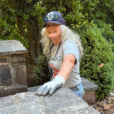 Woman wearing gloves and a hat building or repairing a stone wall outdoors surrounded by greenery.