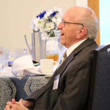 Elderly man in suit and glasses seated at a table with folded hands and floral centerpiece in background