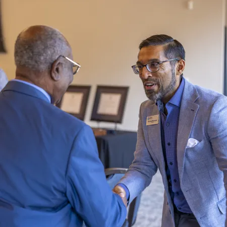 Two professionally dressed men shaking hands during a business event with a man clapping in the background