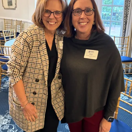 Two smiling women wearing glasses posing indoors at a social event with name tags and elegant decor