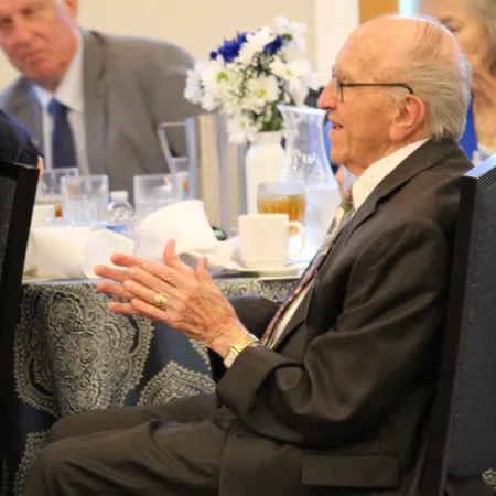 Elderly man in suit with hands clasped sits at a formal event table with flowers and beverages in background.