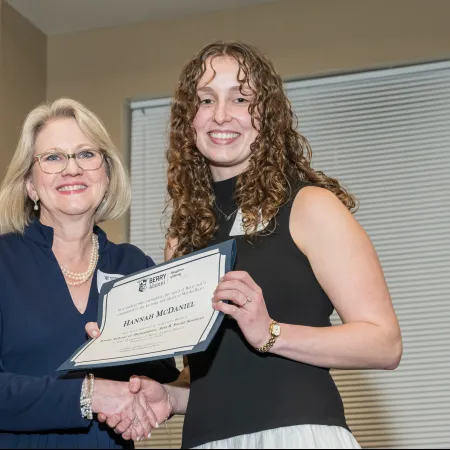 Two women smiling and shaking hands as one receives a certificate of achievement indoors.