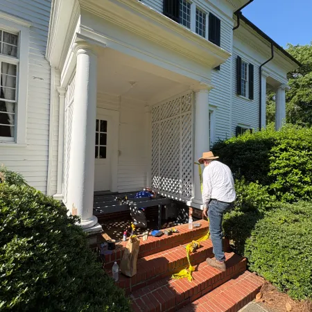Person wearing a hat working on brick steps at the entrance of a white colonial-style house with columns and greenery.