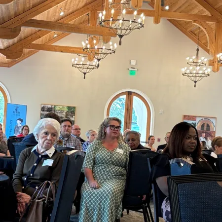 Audience seated attentively in a wooden-beamed room with large arched windows and chandeliers during an indoor event