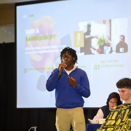 Young man in blue sweater presenting with microphone during a business pitch event with panel judges in background