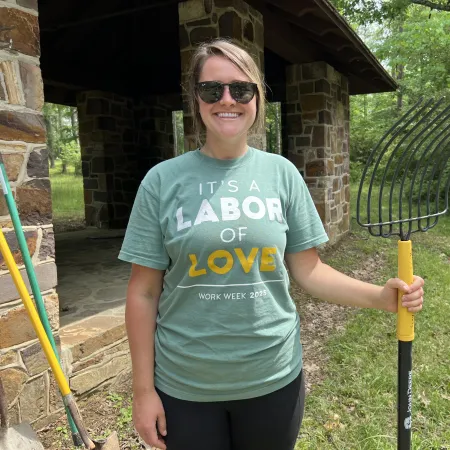 Smiling woman in sunglasses holding a pitchfork wearing a green shirt that says It's a labor of love Work Week 2023