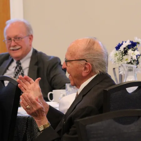 Two elderly men in suits engaged in conversation at a formal indoor event with floral centerpiece on table.