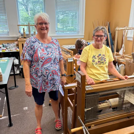 Two elderly women near weaving looms in a bright room with large windows and craft supplies.