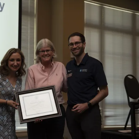 Three people smiling, one holding a framed certificate, standing in a room with a presentation screen in the background.
