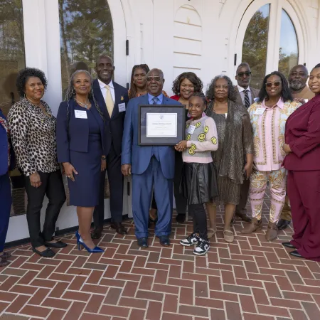 Group of diverse adults and a child posing outdoors with a certificate of recognition in front of white building doors.