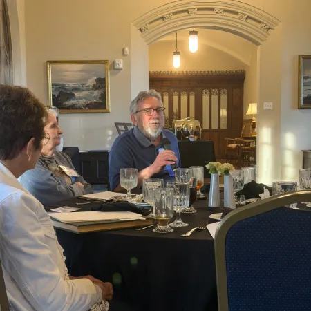 Group of people seated around a table in a formal room, engaged in discussion with glasses and notebooks.