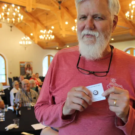 Older man with white beard and glasses holds a card in a warmly lit wood-beamed room with chandeliers.