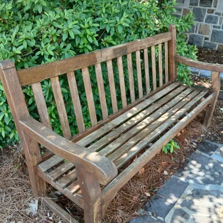 Wooden garden bench next to green bushes and stone pathway in outdoor setting with sunlight and shadows.