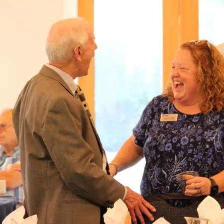 Elderly man and woman laughing and interacting at a formal dining table with white napkins and glassware.