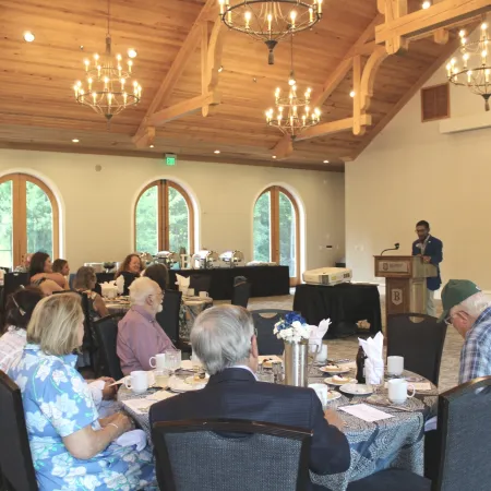 Guests seated at round tables in a banquet hall listening to a speaker at a podium during a daytime event.