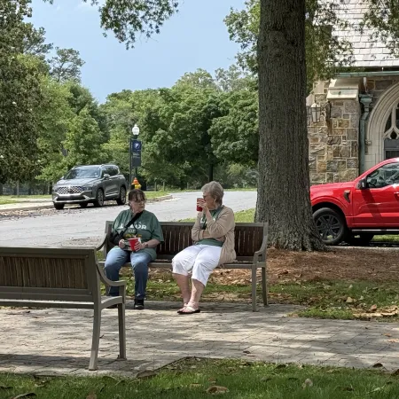 Two women sitting on a bench in a park area near a church, enjoying drinks and talking on a sunny day.