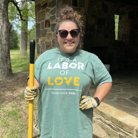 Smiling woman in sunglasses and gloves holding a yellow tool outside a stone cabin, wearing a labor of love t-shirt